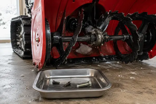 Close-up of a tracked snow blower auger and a parts tray containing broken shear bolts and one replacement bolt.